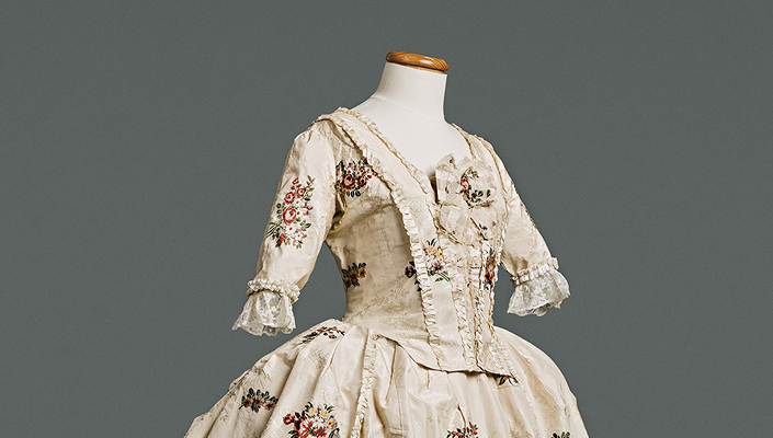 A robe à l’anglaise gown on a mannequin. The top of the gown is shown with a bodice fitted at front and back, and a waistline marked by a seam. It is cream-coloured with embroidered flowers, and frilly lace cuffs.