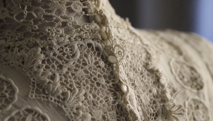 Details of the neckline of a 1930s wedding dress, showing intricate lace embroidery and small ivory-coloured buttons.