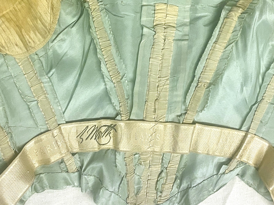 A close-up of the interior bodice of a dress shows several boning channels leading to an interior waistband that bears the branded insignia of Worth.