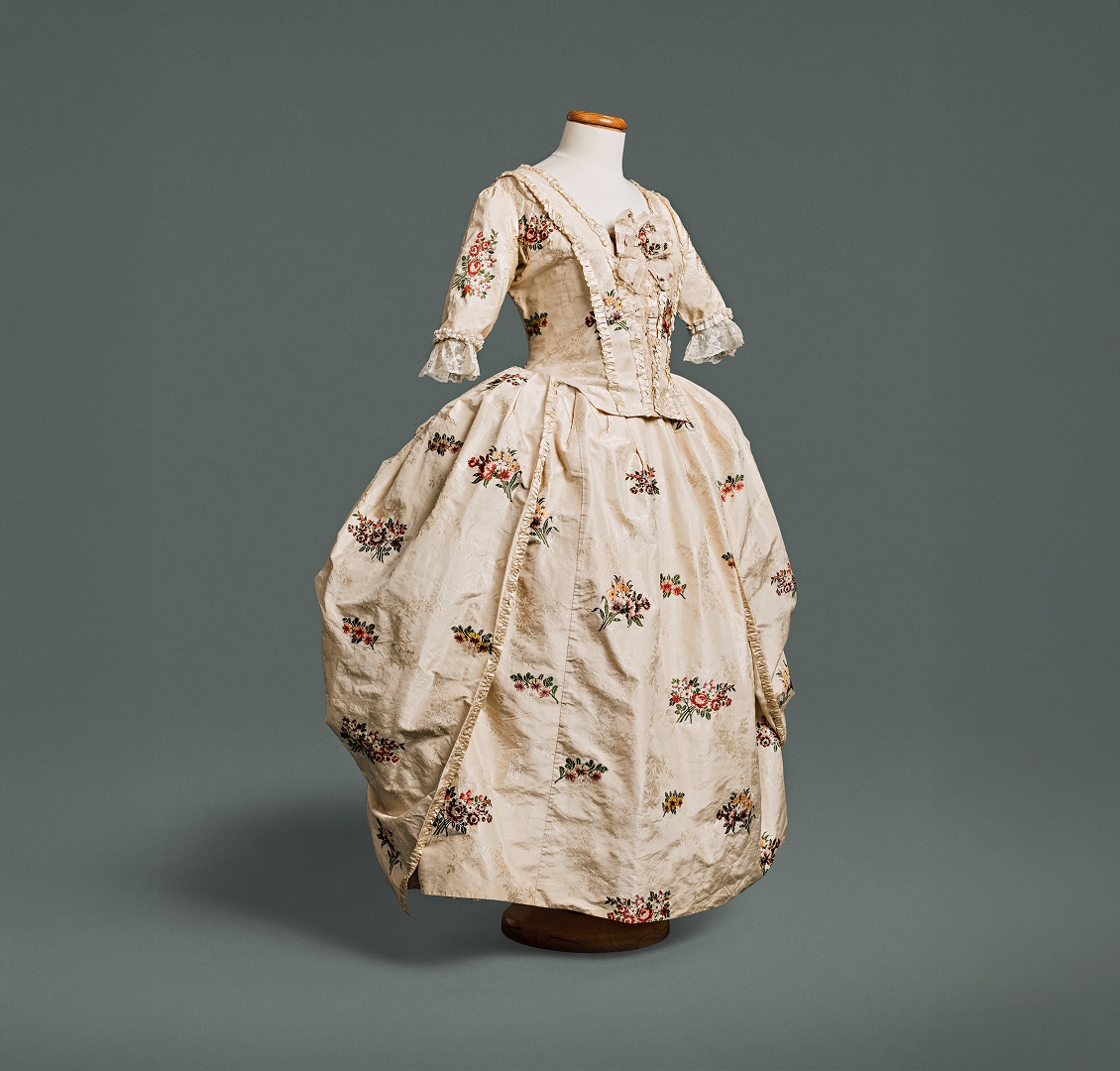 A gown with a bodice fitted at front and back, and a waistline marked by a seam, made of silk decorated with flowers against a white background.