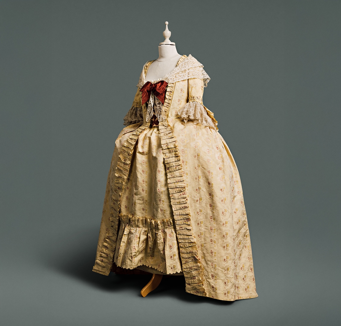 A cream-coloured gown features a loose panel at the back of the bodice, displaying large amounts of expensive cloth patterned with pomegranates, roses, and other flowers against a damask woven pattern of lace bands. A large auburn bow and lace shawl are draped across the top of the gown.