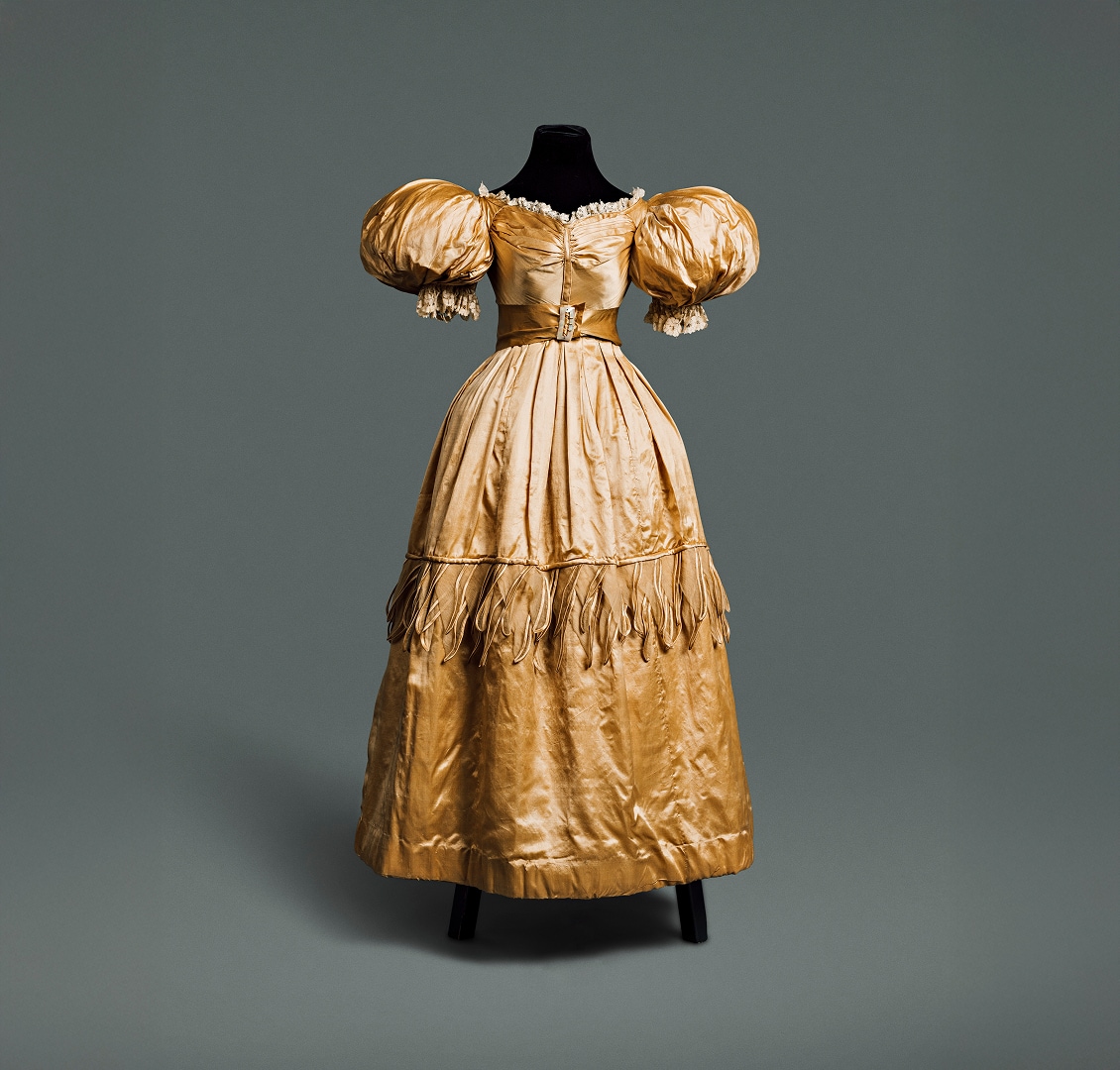 Gold silk gown with long wide skirt, puffy sleeves around the shoulders, and a narrow waistline accented by a belt made of similar material.
