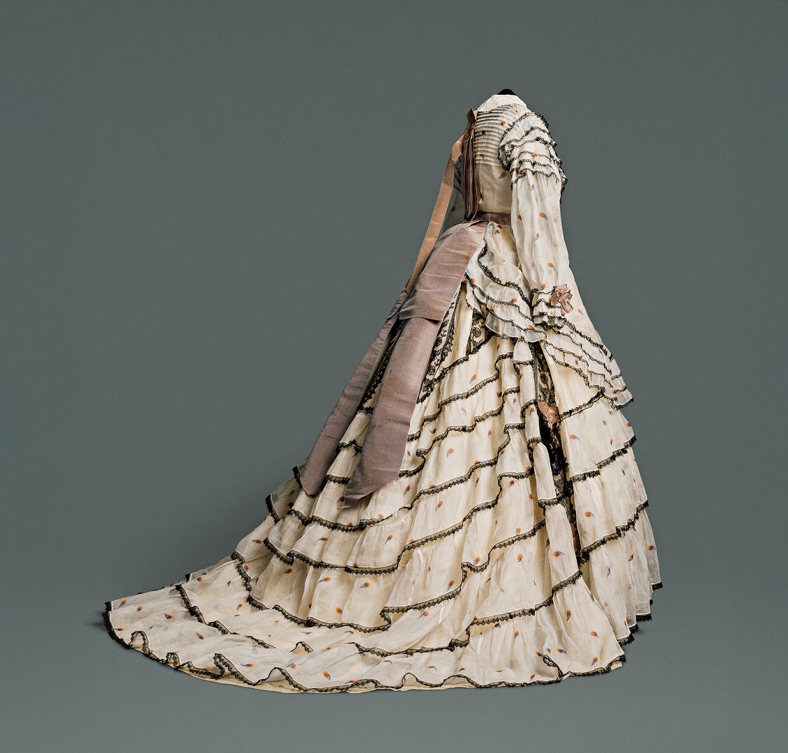 A long, voluminous skirt made of multiple cream-coloured patterned cotton and linen layers accented with black lace. A light brown sash hangs down from the waistline at the back of the dress.