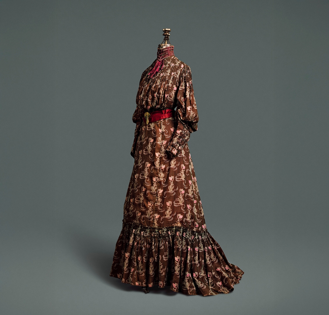 A four-piece dress made of chocolate-brown cotton material patterned with a light brown and pink thistle print. The dress is accented with a deep red silk belt, fastened by a gold clip, and collar.