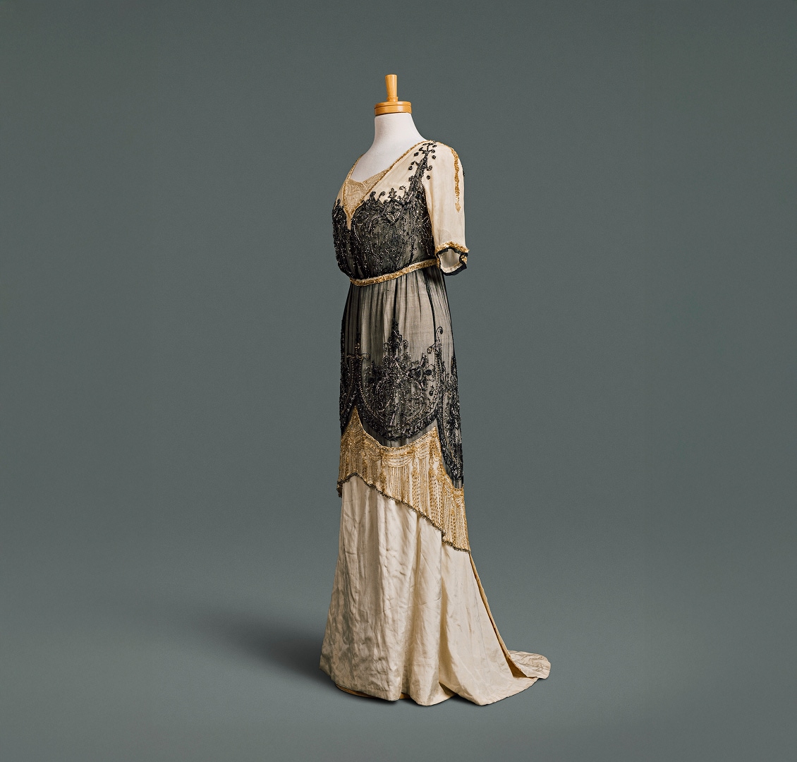 A long, narrow cream-coloured silk dress with low neckline and high waistline, overlayed with black and gold beaded tunic overskirt.