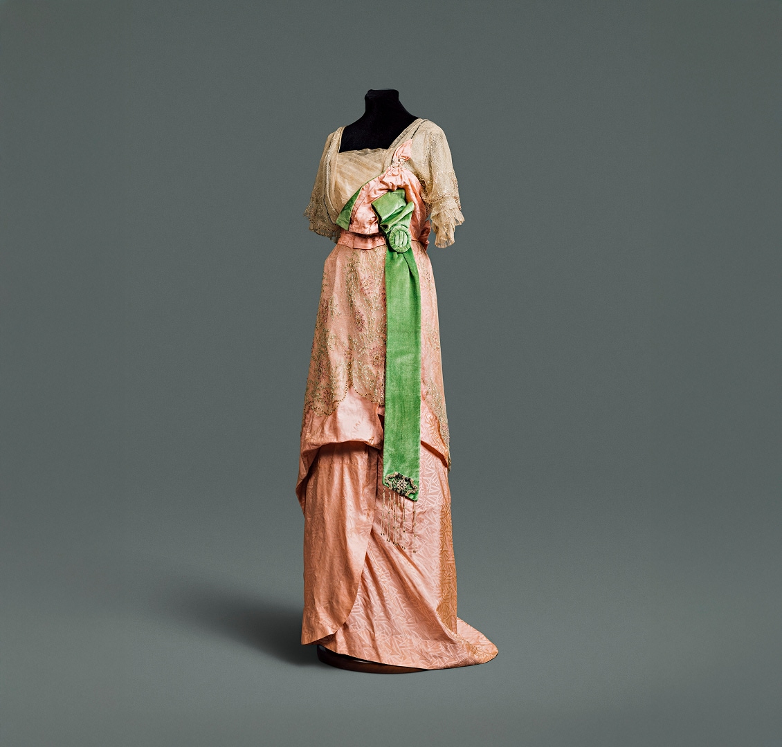 A pink silk dress with draped skirt and wide emerald green sash, covered by an intricate lace bodice and overskirt.