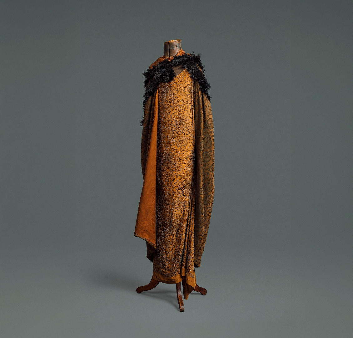 A long, loose dress made of auburn silk covered in a floral pattern made of black beads. Covering it is a matching cape made of heavier patterned material and a black feather boa.