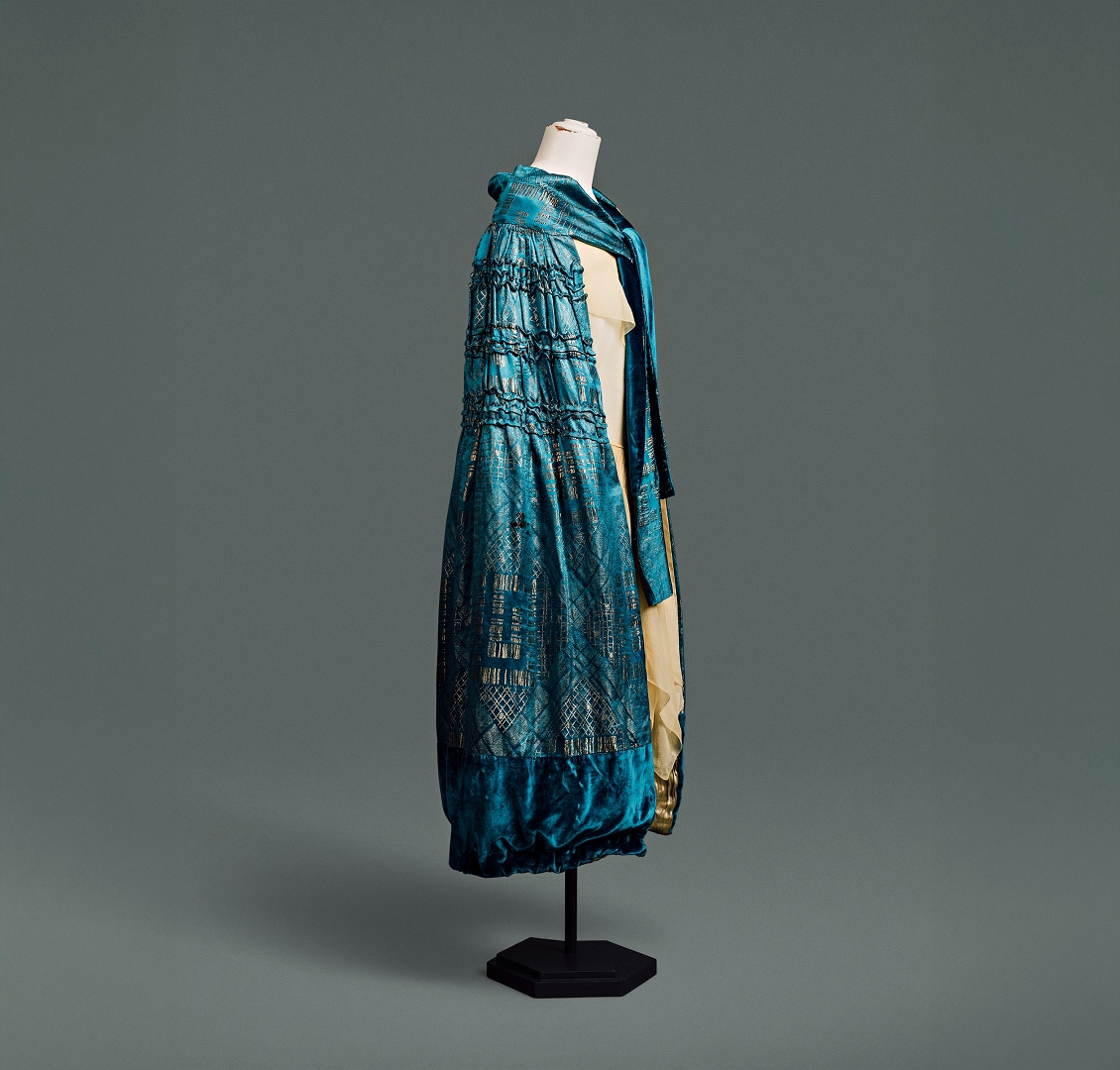 A blue silk cape covered in an Art Deco damask pattern of metallic gold geometric forms, hangs to just below knee length.