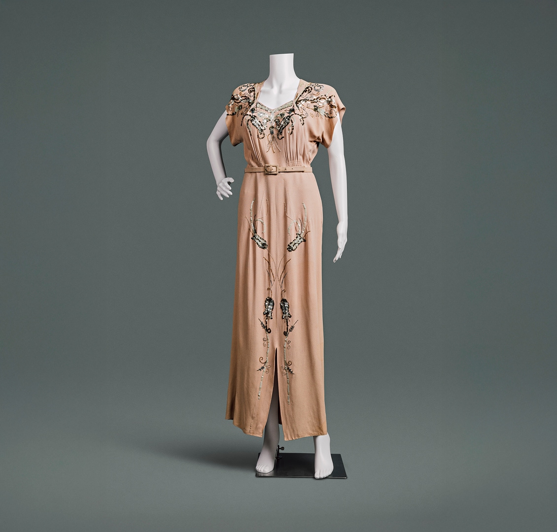 A peach-coloured dress with beaded floral accents, heavily padded shoulders, and a slim skirt. A high waistline is accented by a thin belt made of the same material.