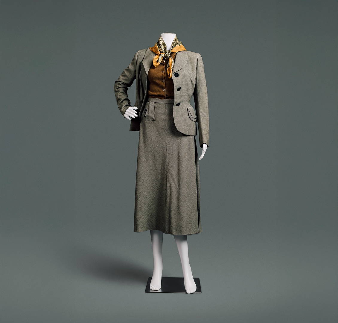 An English grey wool suit with jacket and skirt. The suit features broad, sloping shoulders over an auburn button-up blouse, a tiny waistline, and a longer, ample skirt. A paisley scarf around the neck provides a perfect accent.