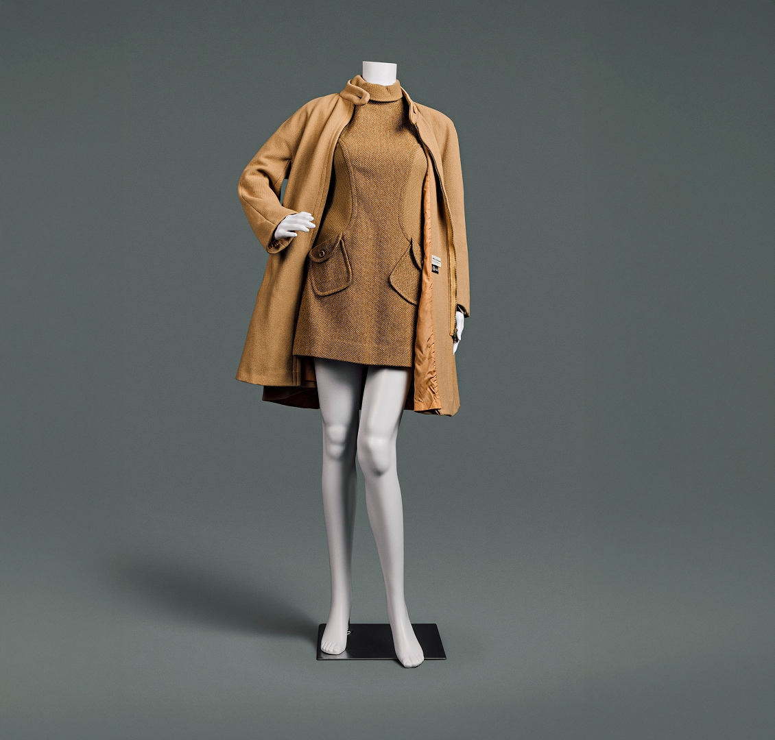 A very short camel-coloured, turtleneck wool dress with herringbone pattern and angled pockets accompanied by heavy camel coat that comes down to just above knee.