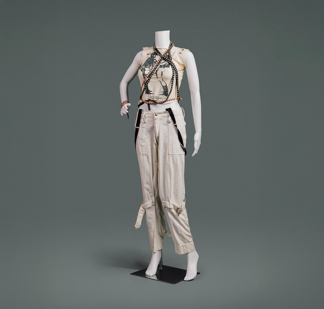 A t-shirt with ripped sleeves and Vivienne Westwood’s iconic “Two Cowboys” print on its front, cream-coloured cotton pants with large pockets and stitched belts that tighten around each leg above the knee, and black and pink studded leather belts draped around the neck overlaying the t-shirt.