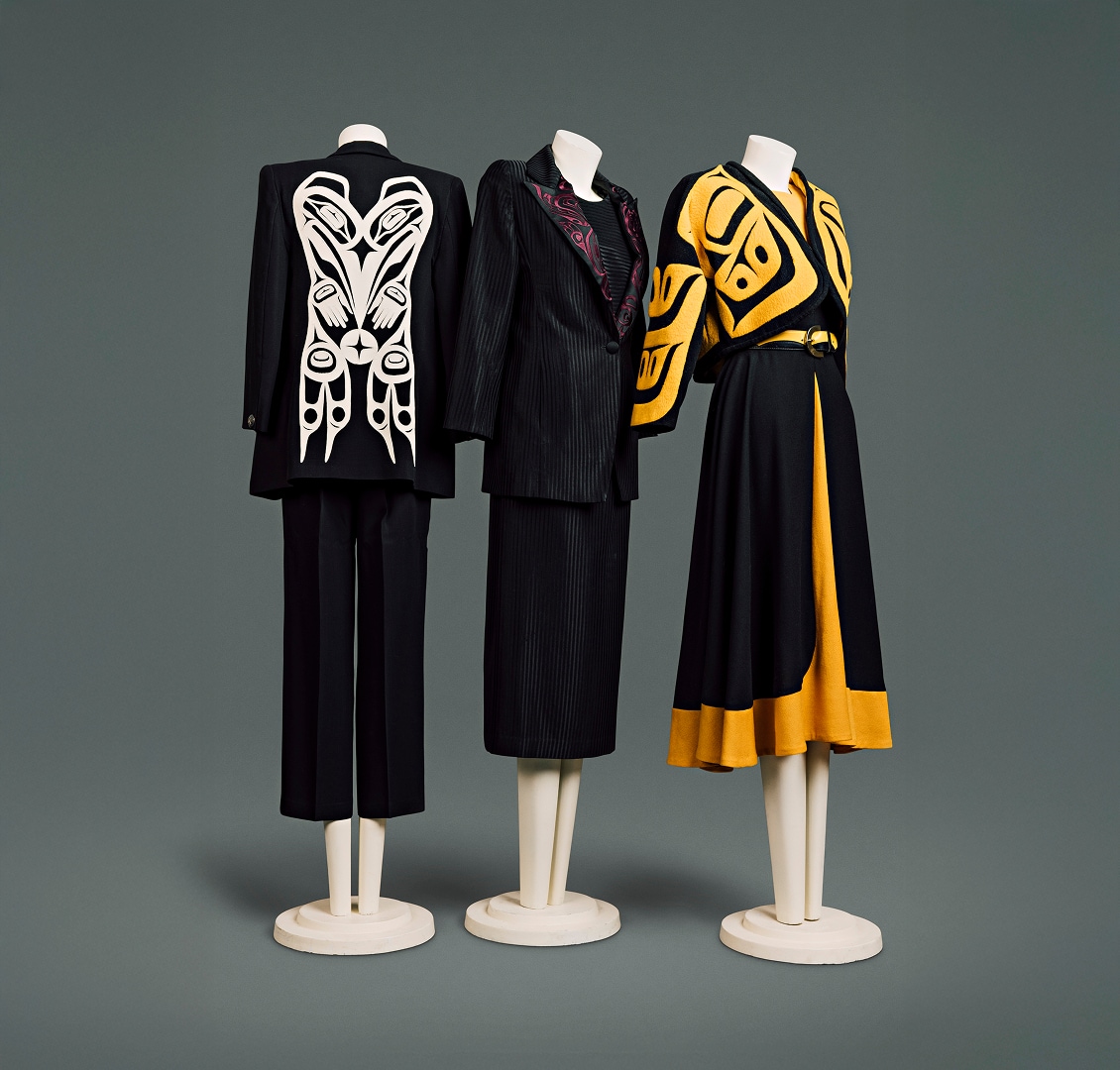 A collection of three contemporary wool suits for women. The first has a black jacket with prominent white Haida motif on the back panel and black pants which come down to mid-calf. The second is made of black pin-striped material with red haida motif on the lapel of the jacket. It is accompanied by matching blouse and a skirt which comes down just below the knee. The third has a shorter, cape-like black jacket, patterned with prominent bright-yellow Haida motif, over a yellow blouse, and yellow-and-black skirt which comes down just below the knee. It is accented by a black-and-yellow leather belt with simple crescent-shaped buckle.