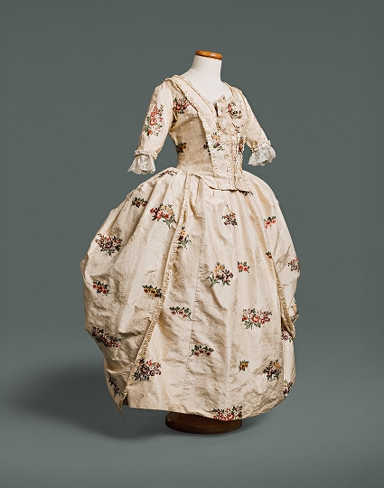 A gown with a bodice fitted at front and back, and a waistline marked by a seam, made of silk decorated with flowers against a white background.