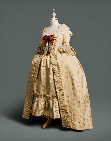 A cream-coloured gown features a loose panel at the back of the bodice, displaying large amounts of expensive cloth patterned with pomegranates, roses, and other flowers against a damask woven pattern of lace bands. A large auburn bow and lace shawl are draped across the top of the gown.