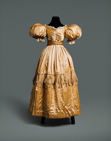 Gold silk gown with long wide skirt, puffy sleeves around the shoulders, and a narrow waistline accented by a belt made of similar material.