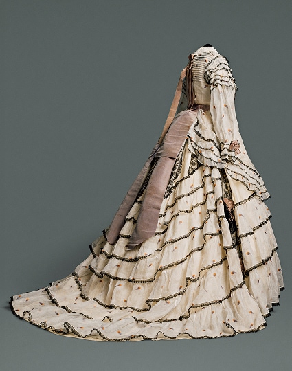 A long, voluminous skirt made of multiple cream-coloured patterned cotton and linen layers accented with black lace. A light brown sash hangs down from the waistline at the back of the dress.