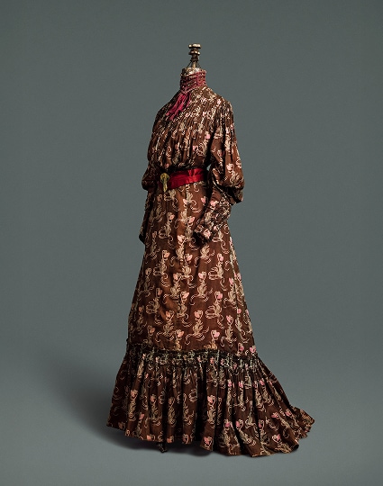 A four-piece dress made of chocolate-brown cotton material patterned with a light brown and pink thistle print. The dress is accented with a deep red silk belt, fastened by a gold clip, and collar.