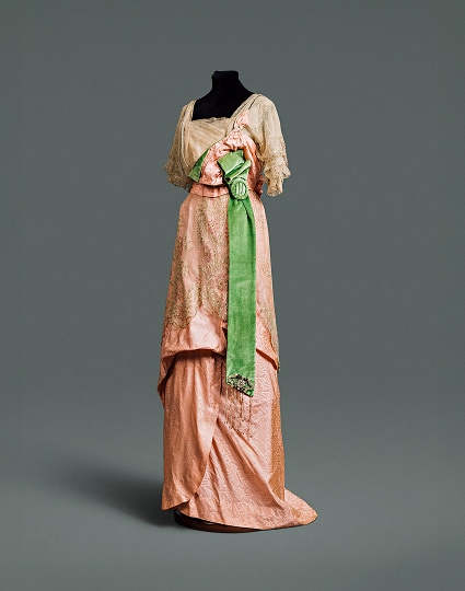 A pink silk dress with draped skirt and wide emerald green sash, covered by an intricate lace bodice and overskirt.
