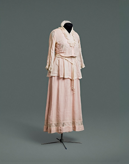 A simple pink cotton one-piece dress designed to appear to be a suit composed of a jacket over a blouse, worn with a matching skirt.