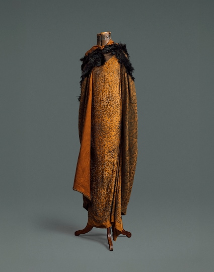 A long, loose dress made of auburn silk covered in a floral pattern made of black beads. Covering it is a matching cape made of heavier patterned material and a black feather boa.
