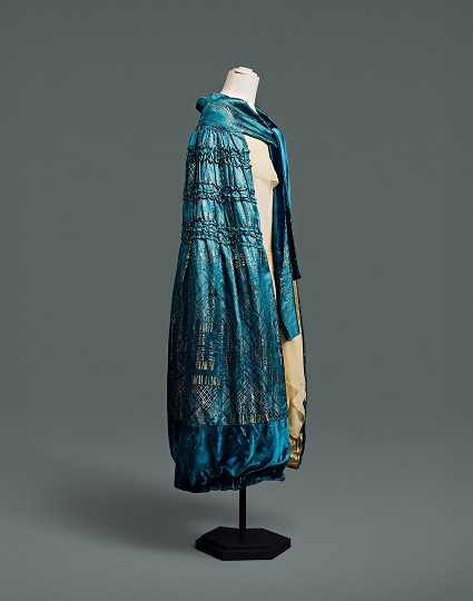 A blue silk cape covered in an Art Deco damask pattern of metallic gold geometric forms, hangs to just below knee length.