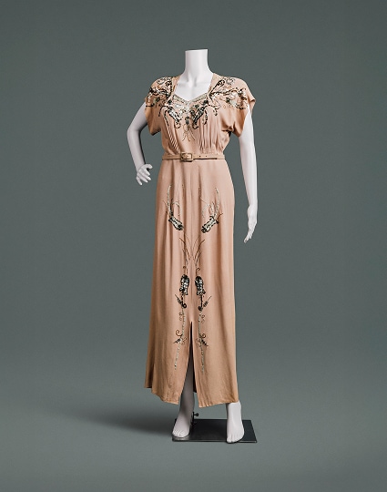 A peach-coloured dress with beaded floral accents, heavily padded shoulders, and a slim skirt. A high waistline is accented by a thin belt made of the same material.