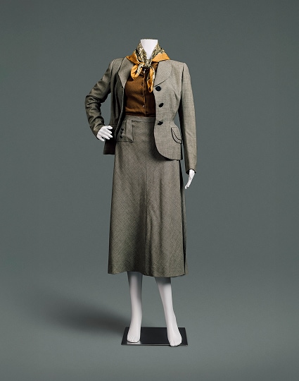 An English grey wool suit with jacket and skirt. The suit features broad, sloping shoulders over an auburn button-up blouse, a tiny waistline, and a longer, ample skirt. A paisley scarf around the neck provides a perfect accent.