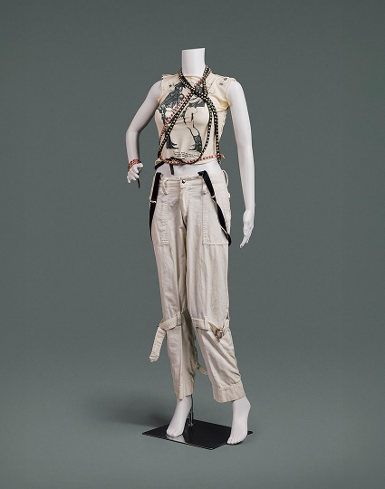 A t-shirt with ripped sleeves and Vivienne Westwood’s iconic “Two Cowboys” print on its front, cream-coloured cotton pants with large pockets and stitched belts that tighten around each leg above the knee, and black and pink studded leather belts draped around the neck overlaying the t-shirt.