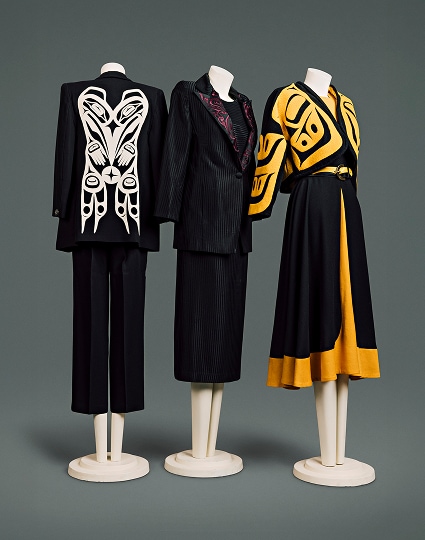 A collection of three contemporary wool suits for women. The first has a black jacket with prominent white Haida motif on the back panel and black pants which come down to mid-calf. The second is made of black pin-striped material with red haida motif on the lapel of the jacket. It is accompanied by matching blouse and a skirt which comes down just below the knee. The third has a shorter, cape-like black jacket, patterned with prominent bright-yellow Haida motif, over a yellow blouse, and yellow-and-black skirt which comes down just below the knee. It is accented by a black-and-yellow leather belt with simple crescent-shaped buckle.