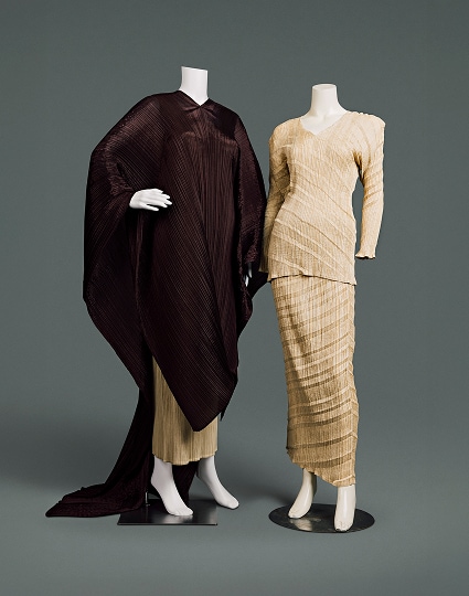 Two contemporary ensembles made of polyester. The first consists of a form-fitting, tan-coloured blouse and full length skirt, whose material is crinkled vertically and striped with horizontal diagonal shades. The second consists of a similar skirt mostly covered by a rich brown tunic blanketing the arms and body, extending below the knees in the front and trailing onto the floor like a long cape at the back.