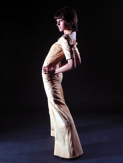 A model from the 1970s wearing a one-piece pant suit poses with arched back and hands on her hips.