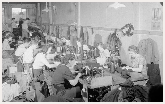 A large group of women sit at both sides of a long table assembling garments with sewing machines.