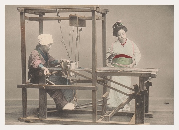 A Japanese women sits operating a loom while another feeds thread into the machine. Both wear traditional kimono.