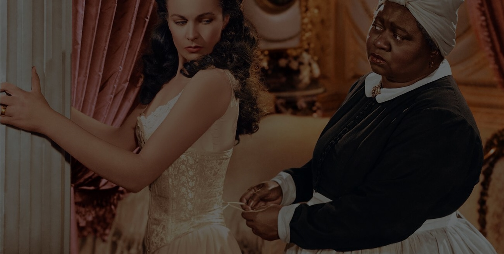 A production still from Gone with the Wind shows Mammy tightening Scarlett’s corset while she steadies herself against a column.