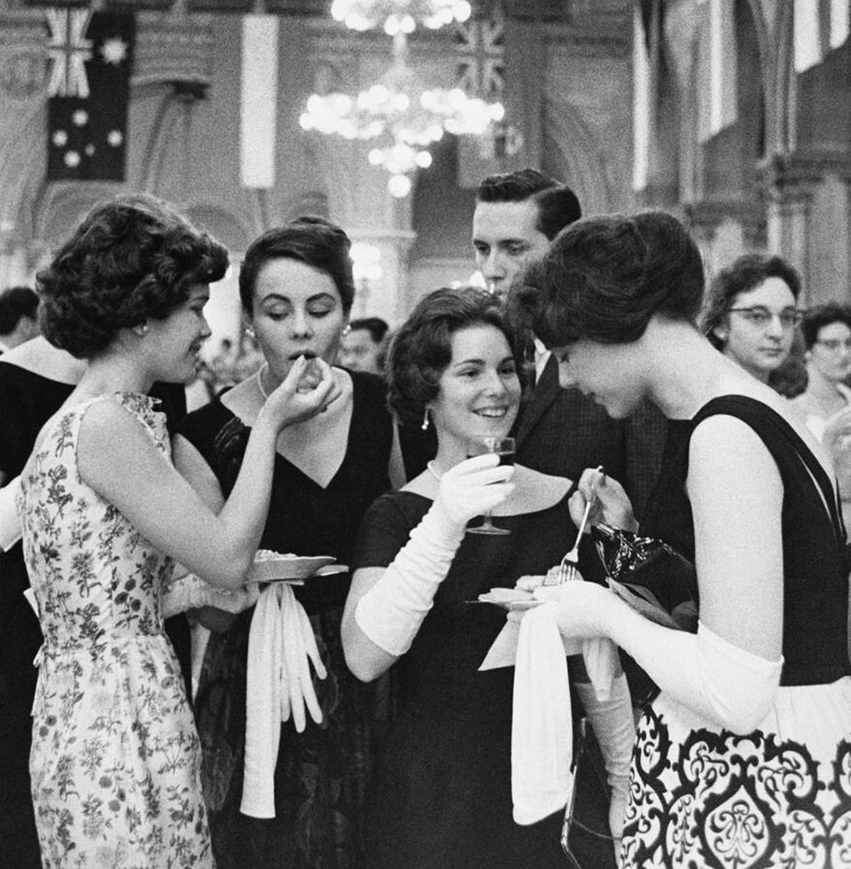 Four women, wearing long white gloves, eat cake and drink wine at an upscale cocktail party.