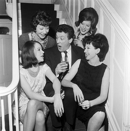 An eligible bachelor sits on the stairs with a drink, surrounded by four beautiful women.