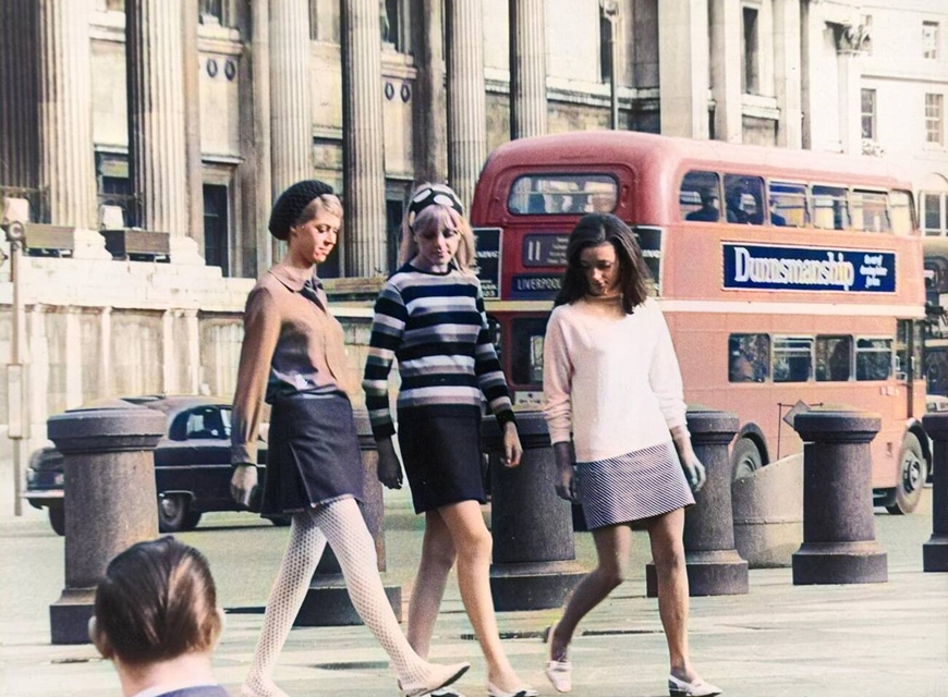 Three young women wearing mini skirts walk through Trafalgar Square with a double decker bus in the background.