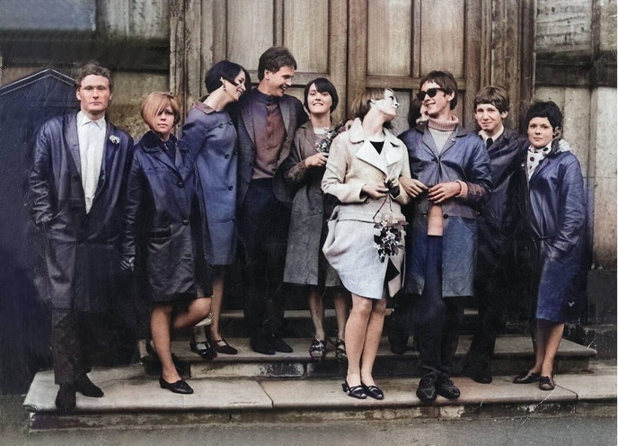 A group of stylish mod couples wearing long coats stands on the stairs at the entrance of an old building.