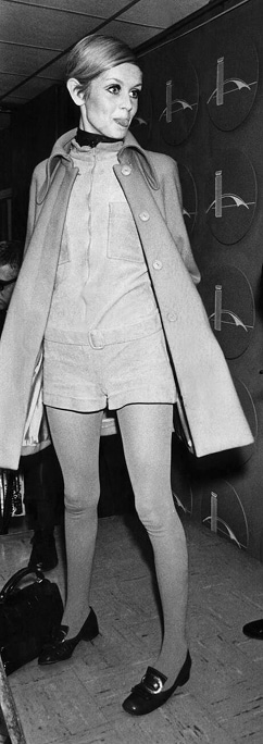 Twiggy wearing a one-piece romper and pea coat, sticks her tongue out at photographers.