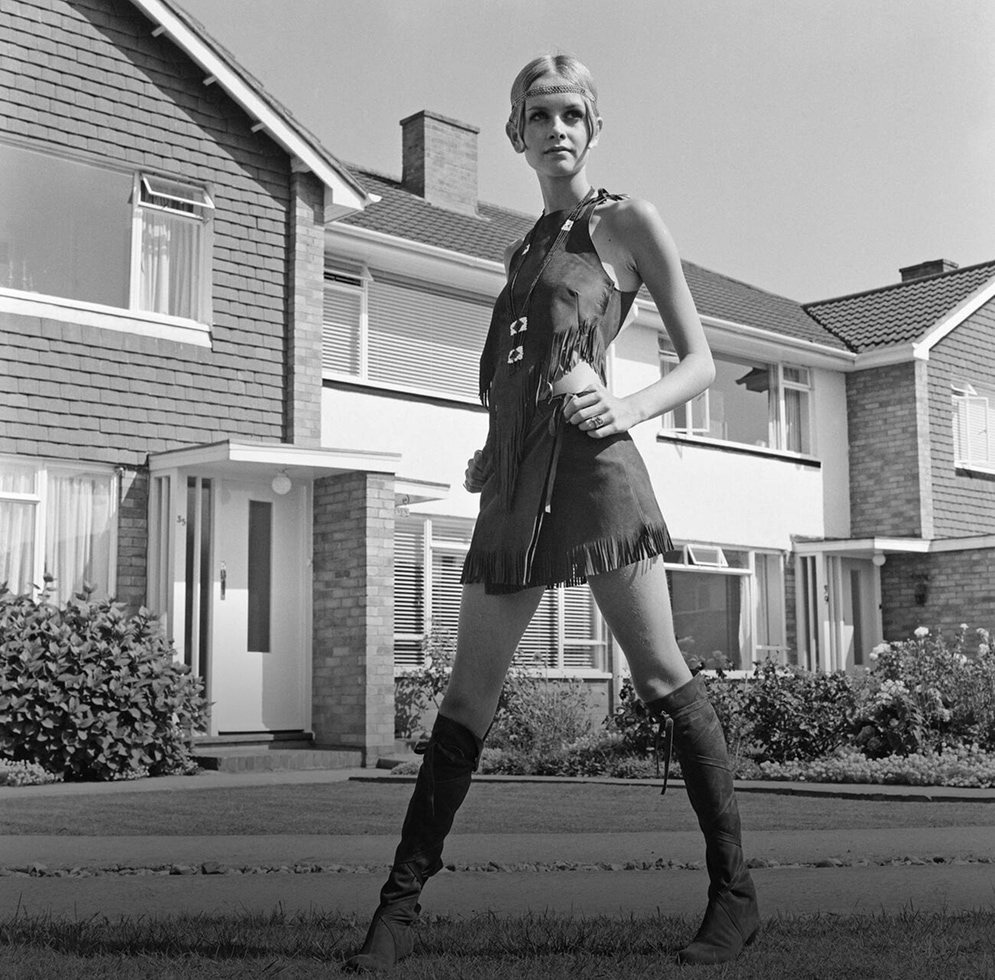 Twiggy, in fringed leather mini dress and matching boots, strikes a pose in front of suburban homes.
