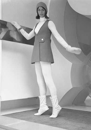 The model wearing a sleeveless mini-dress in bright green wool, orange wool bonnet, and white mittens and buttoned boots.
