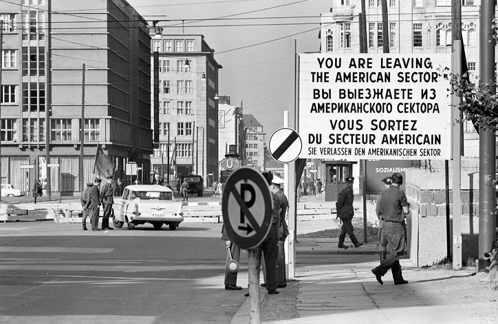 Guards stop a car at a checkpoint in West Berlin. A sign in the foreground reads “You are leaving the American Sector.”