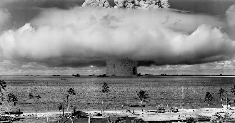 A giant mushroom cloud over a Pacific atoll. Ships surround the explosion, while a beach and palm trees are visible in the immediate foreground.